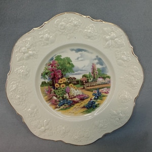May include: A white decorative plate with a scalloped edge and embossed floral designs. The center features a colorful garden scene with flowers, trees, and a pathway. The rim has a gold trim.