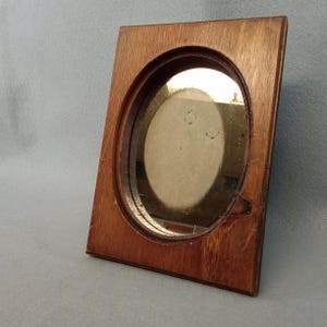 May include: A rectangular wooden picture frame with a prominent oval-shaped cutout in the center. The frame features a warm, medium-brown wood grain finish with visible natural textures and a small knot detail on the lower right side of the oval.