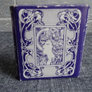 May include: A purple box with a white design featuring a stylized image of a woman's face. The design is framed by a decorative border.