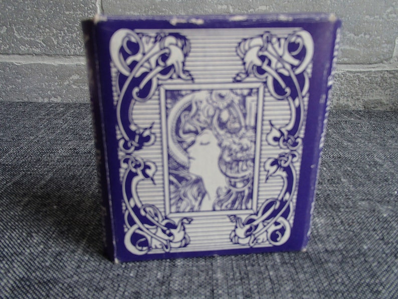 May include: A purple box with a white design featuring a stylized image of a woman's face. The design is framed by a decorative border.