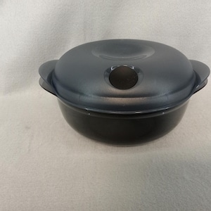 May include: A black, round food storage container with a matching lid. The lid has a dark gray hue and a black handle. The container has two side handles for easy carrying. The container is sitting on a white surface.