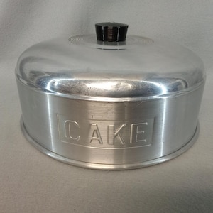 May include: A vintage aluminum cake carrier with a domed lid and a black handle. The word "CAKE" is embossed on the side. The carrier is silver and has a circular design on the lid.