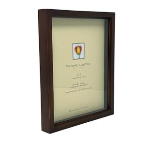 May include: A dark brown wooden picture frame with a glass front. The frame is designed to hold an 8x10 inch photo or a small keepsake. The frame has a small red, orange, and yellow flower design in the top right corner. The text "THE DANIELS COLLECTION" is printed in black at the top of the frame. The text "2-in-1 treasure box home" is printed in black below the flower design. The text "holds a 8 x 10 image or display favorite keepsakes" is printed in black below the text "2-in-1 treasure box home". The text "move one easy piece for the frame you want" is printed in black below the text "holds a 8 x 10 image or display favorite keepsakes". The text "DENNIS DANIELS COMPANY" is printed in black at the bottom of the frame.