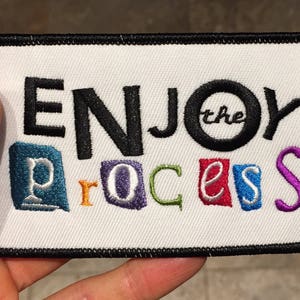 May include: Embroidered patch with the words "ENJOY the PROCESS" in black, and colorful letters. The patch has a white background with a black border. The word "the" is inside a black circle.