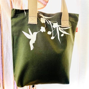 May include: A green canvas tote bag with a white hummingbird and floral design. The bag has tan straps.