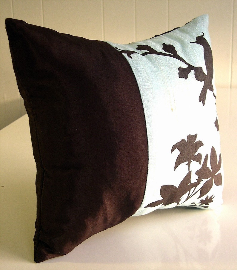 K&ouml;nnte beinhalten: Ein braunes und hellblaues Kissen mit Blumendesign. Das Kissen hat eine braune Seite und eine hellblaue Seite mit einem schwarzen Blumendesign.