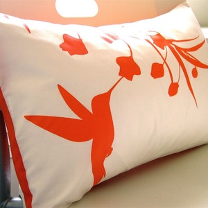 May include: A white pillow with an orange silhouette of a hummingbird and flowers. The pillow is made of a soft, white fabric and has a subtle sheen.