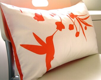 May include: A white pillow with an orange silhouette of a hummingbird and flowers. The pillow is made of a soft, white fabric and has a subtle sheen.