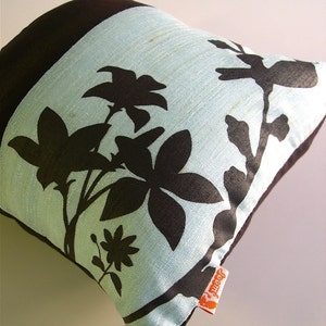 K&ouml;nnte beinhalten: Ein hellblaues Kissen mit einem schwarzen Blumendesign. Das Kissen hat eine braune R&uuml;ckseite und ein kleines orangefarbenes Etikett mit der Aufschrift "Jeannie".