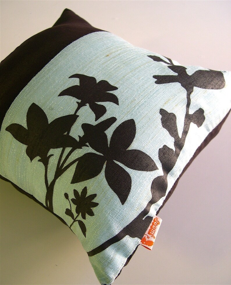 K&ouml;nnte beinhalten: Ein hellblaues Kissen mit einem schwarzen Blumendesign. Das Kissen hat eine braune R&uuml;ckseite und ein kleines orangefarbenes Etikett mit der Aufschrift "Jeannie".