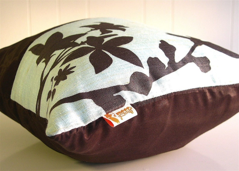 K&ouml;nnte beinhalten: Ein braunes und hellblaues Kissen mit einem schwarzen Blumendesign. Das Kissen hat eine braune Stoffr&uuml;ckseite und ein wei&szlig;es Etikett mit dem aufgedruckten Wort "Joomi".