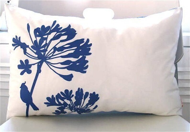 May include: White pillow with a blue silhouette of a flower and a bird. The flower has a large, round head with many petals. The bird is perched on a branch of the flower.