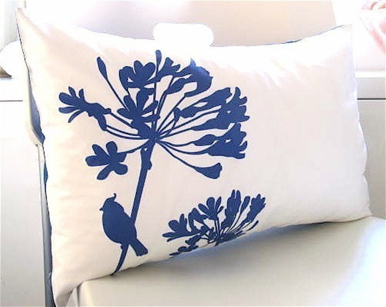 May include: White pillow with a blue silhouette of a flower and a bird. The flower has a long stem and many petals. The bird is perched on the stem.