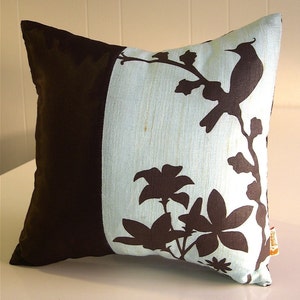 K&ouml;nnte beinhalten: Ein Dekokissen mit braunem und blauem Design. Das Kissen zeigt eine Silhouette eines Vogels und Blumen auf hellblauem Hintergrund. Die andere Seite des Kissens ist einfarbig braun.