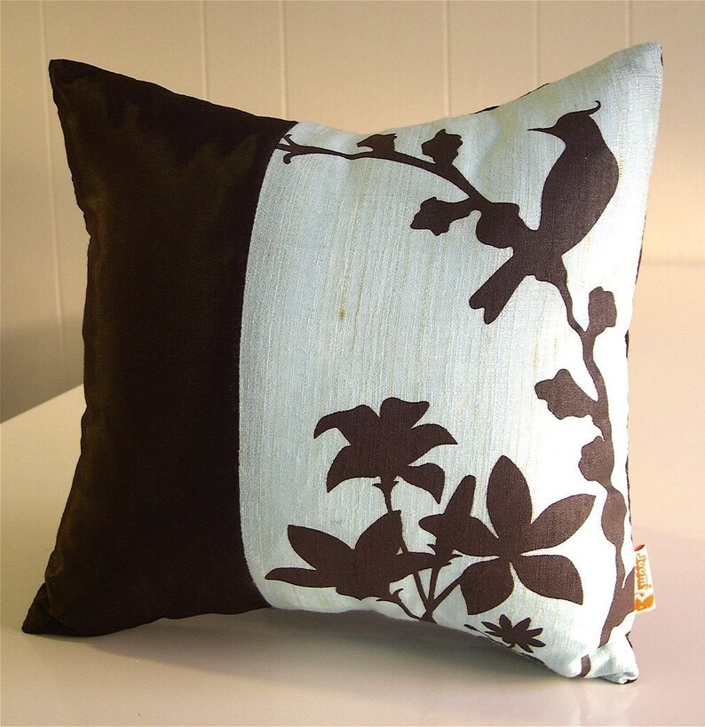K&ouml;nnte beinhalten: Ein Dekokissen mit braunem und blauem Design. Das Kissen zeigt eine Silhouette eines Vogels und Blumen auf hellblauem Hintergrund. Die andere Seite des Kissens ist einfarbig braun.