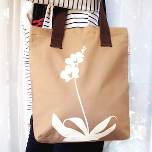 Op de afbeelding: Een bruine canvas tote bag met een wit orchidee&euml;nbloemdessin. De tas heeft bruine hengsels en een witte achtergrond.