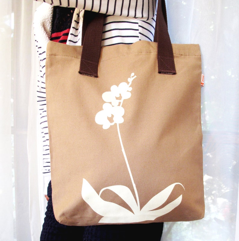 Op de afbeelding: Een bruine canvas tote bag met een wit orchidee&euml;nbloemdessin. De tas heeft bruine hengsels en een witte achtergrond.