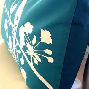 May include: Teal blue pillow with a white silhouette of a flowering branch. The pillow has a red label with the word "Jenny" on it.