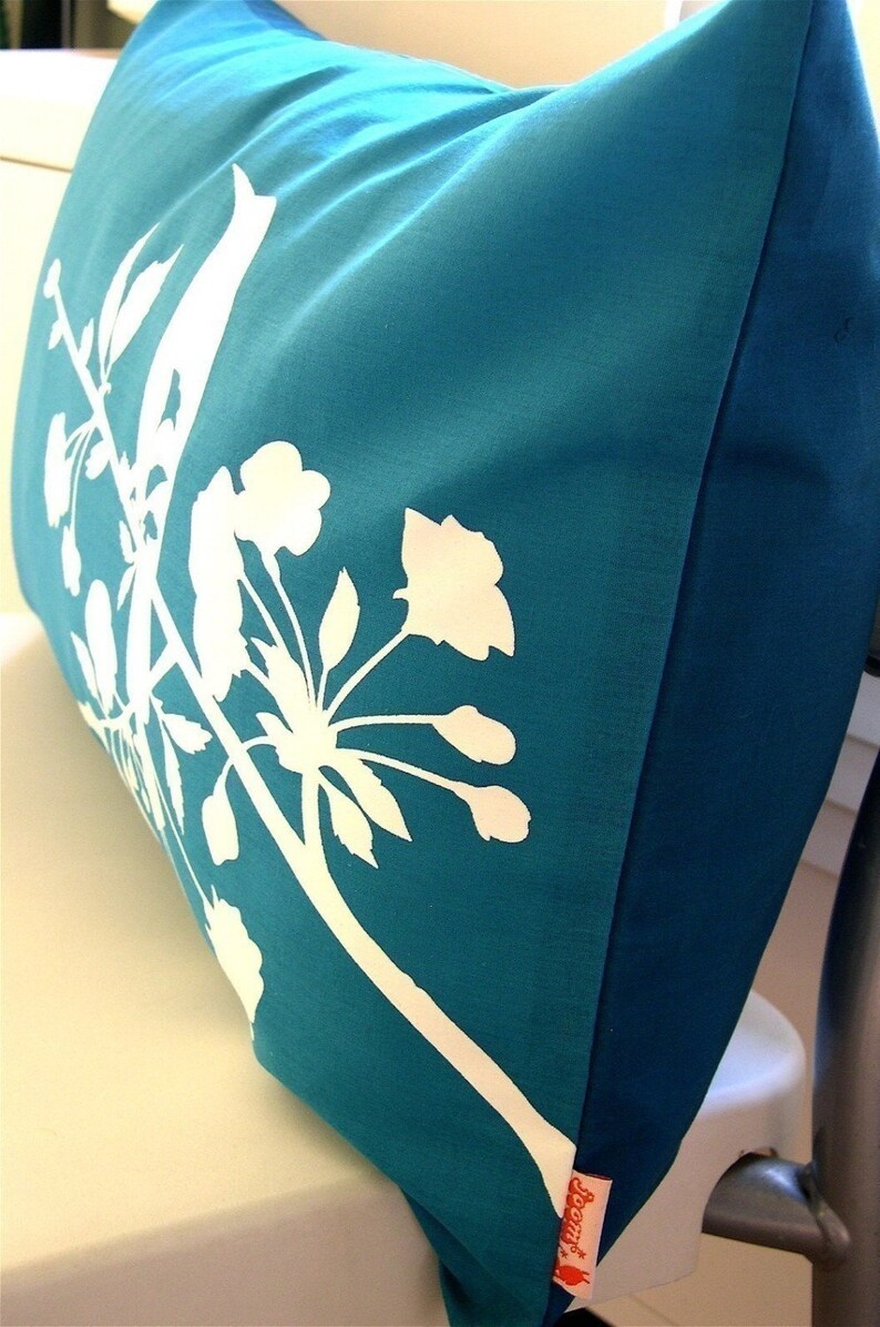 May include: Teal blue pillow with a white silhouette of a flowering branch. The pillow has a red label with the word "Jenny" on it.
