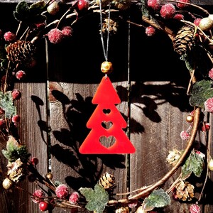 May include: Red Christmas tree ornament with three hearts cut out. The ornament is hanging from a wreath made of evergreen branches, red berries, and gold accents.