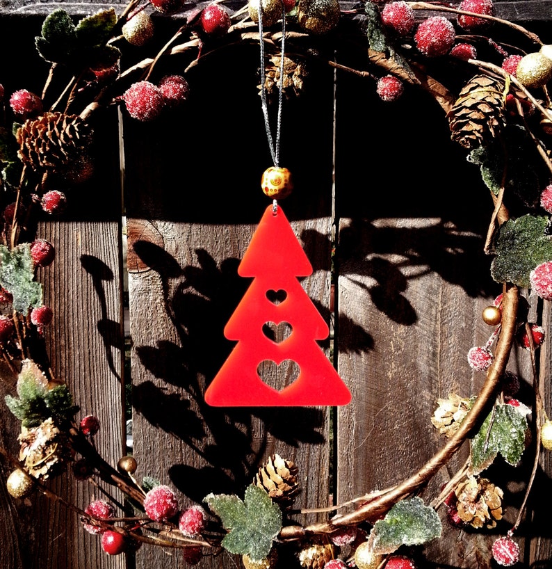 May include: Red Christmas tree ornament with three hearts cut out. The ornament is hanging from a wreath made of evergreen branches, red berries, and gold accents.