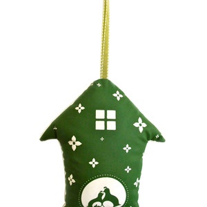 May include: A green fabric house-shaped ornament with a white window and floral pattern. The ornament has a white silhouette of two birds inside a circle at the bottom. The ornament has a green ribbon for hanging.