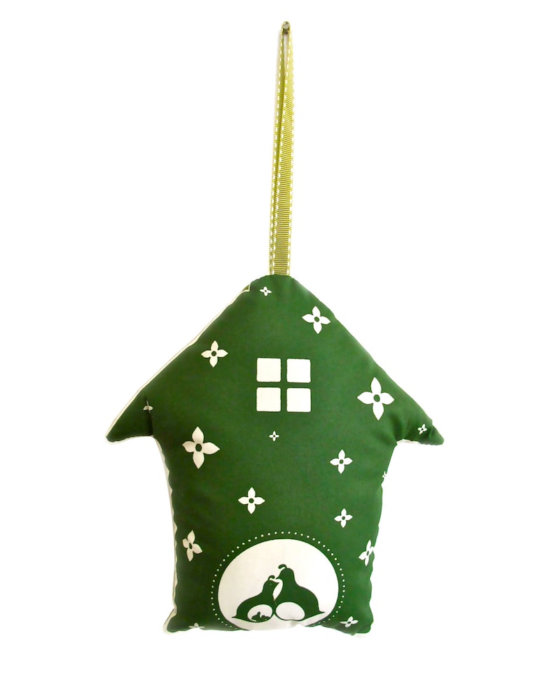 May include: A green fabric house-shaped ornament with a white window and floral pattern. The ornament has a white silhouette of two birds inside a circle at the bottom. The ornament has a green ribbon for hanging.