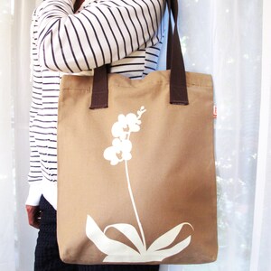 Op de afbeelding: Een bruine canvas tote bag met een wit orchidee&euml;nontwerp. De tas heeft bruine hengsels en een witte achtergrond.