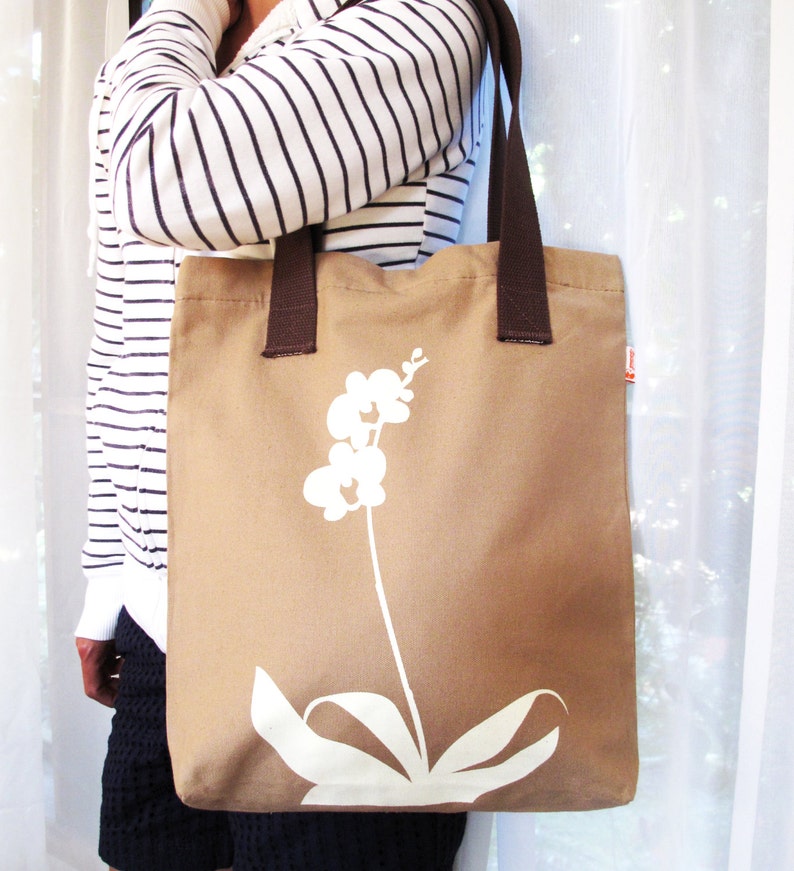 Op de afbeelding: Een bruine canvas tote bag met een wit orchidee&euml;nontwerp. De tas heeft bruine hengsels en een witte achtergrond.