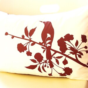 May include: A white pillow with a red silhouette of a bird perched on a branch with flowers. The pillow is made of a soft, white fabric and has a red border.