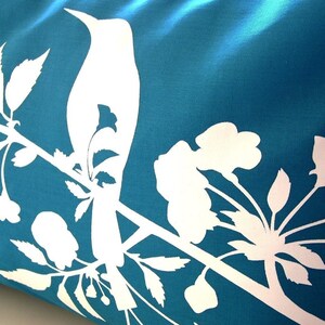 May include: A teal blue pillow with a white silhouette of a bird perched on a branch with white flowers and leaves.