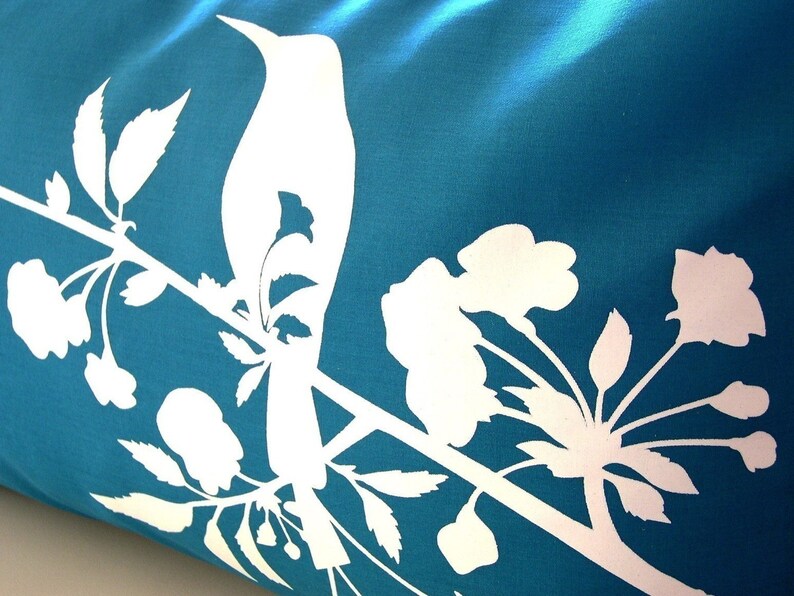 May include: A teal blue pillow with a white silhouette of a bird perched on a branch with white flowers and leaves.
