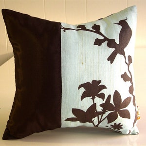 K&ouml;nnte beinhalten: Ein Dekokissen mit braunem und hellblauem Design. Das Kissen zeigt eine Silhouette eines Vogels und Blumen auf hellblauem Hintergrund. Die andere H&auml;lfte des Kissens ist einfarbig braun.