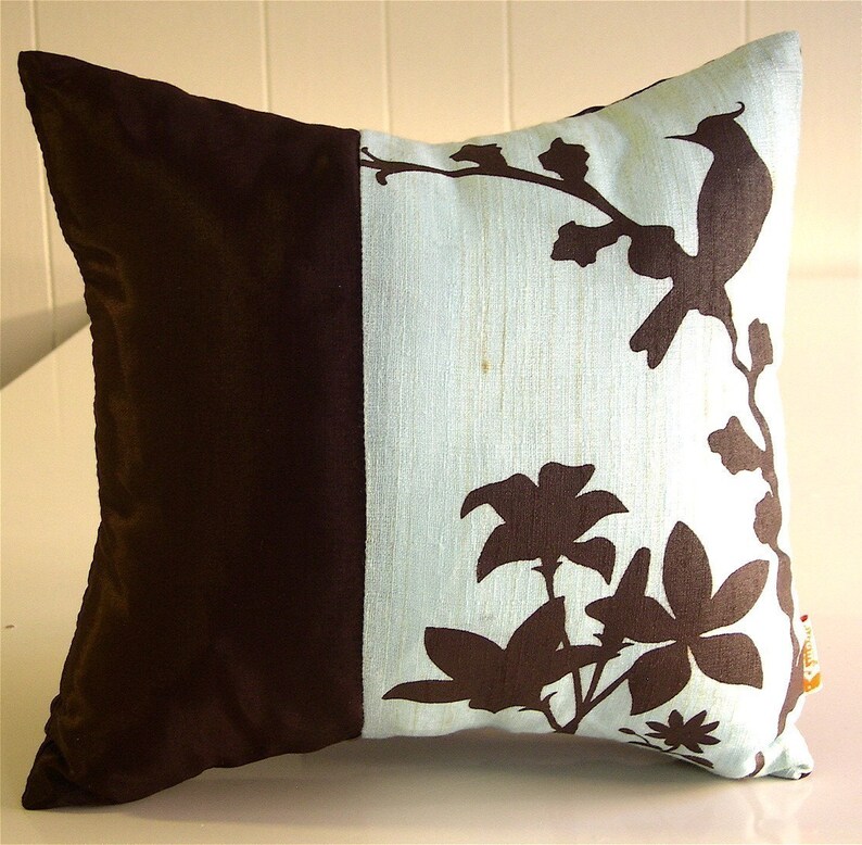 K&ouml;nnte beinhalten: Ein Dekokissen mit braunem und hellblauem Design. Das Kissen zeigt eine Silhouette eines Vogels und Blumen auf hellblauem Hintergrund. Die andere H&auml;lfte des Kissens ist einfarbig braun.