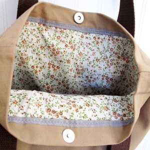 Op de afbeelding: Een beige canvas tote bag met bruine hengsels en een voering met bloemenprint. De tas heeft twee metalen drukknopen aan de bovenkant.