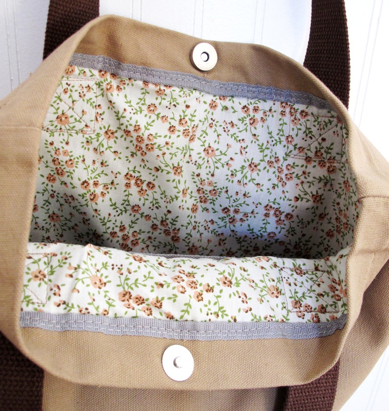 Op de afbeelding: Een beige canvas tote bag met bruine hengsels en een voering met bloemenprint. De tas heeft twee metalen drukknopen aan de bovenkant.