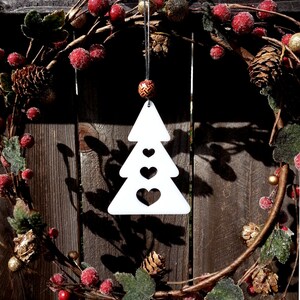 May include: White wooden Christmas tree ornament with three cut-out hearts. The ornament is hanging from a red and gold beaded string on a brown wooden background with a Christmas wreath.