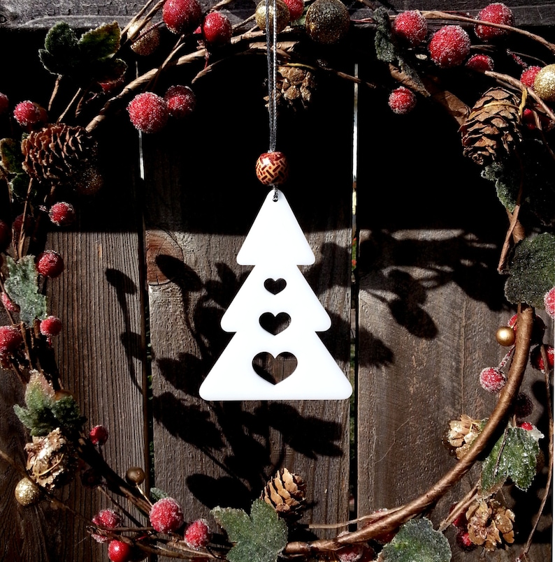 May include: White wooden Christmas tree ornament with three cut-out hearts. The ornament is hanging from a red and gold beaded string on a brown wooden background with a Christmas wreath.