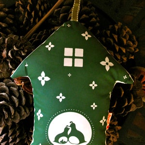 May include: A green fabric house-shaped pillow with white floral designs and a white silhouette of two birds inside a circle. The pillow is sitting on a bed of pine cones.