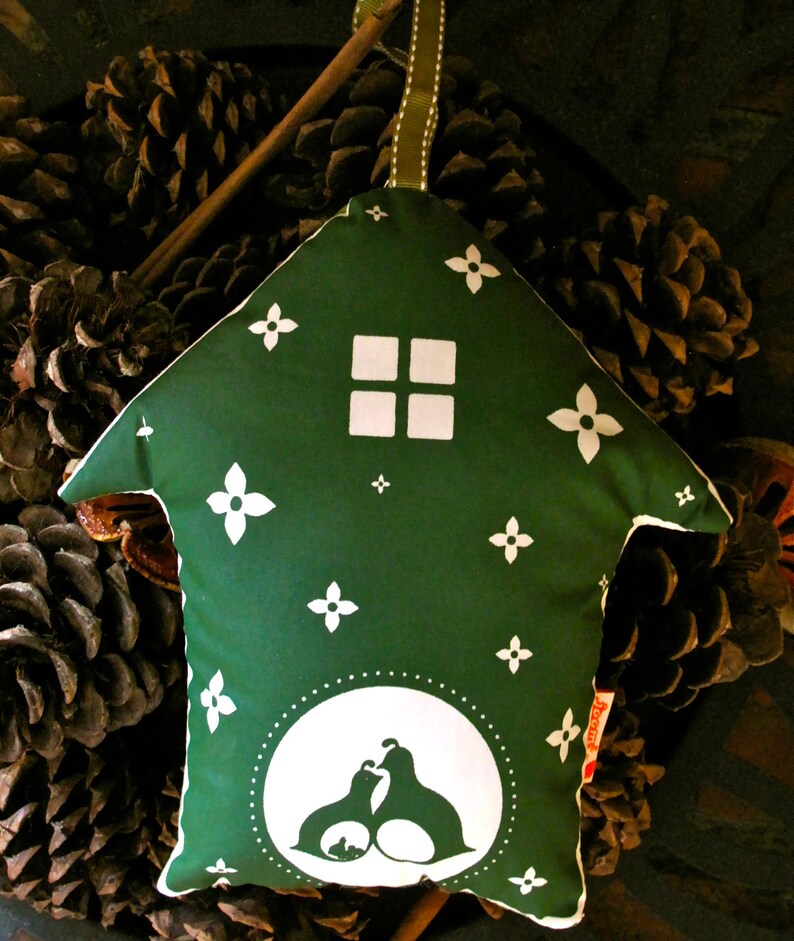 May include: A green fabric house-shaped pillow with white floral designs and a white silhouette of two birds inside a circle. The pillow is sitting on a bed of pine cones.