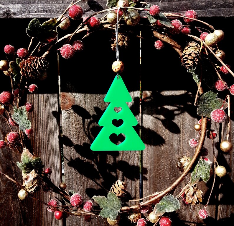 May include: A green Christmas tree ornament with three hearts cut out. The ornament is hanging from a wreath made of green leaves, red berries, and gold accents.
