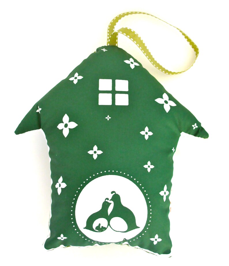 May include: A green fabric house-shaped ornament with a white window and a white silhouette of two birds and a baby bird inside a circle. The ornament has a green and white ribbon loop for hanging.