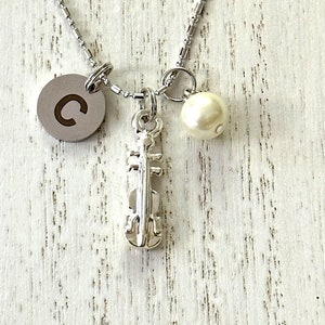 Puede incluir: Collar plateado con un dije de violín, una perla y un colgante circular con la letra "C". La cadena es delicada. Una pieza encantadora para los amantes de la música.