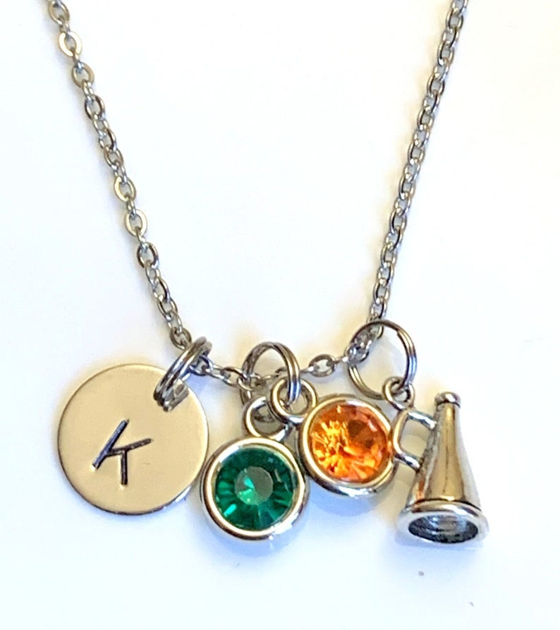 May include: Silver-toned necklace with a chain and multiple charms. Charms include a round pendant with the letter "K", a green and an orange gemstone, and a small megaphone. The necklace is designed for everyday wear.