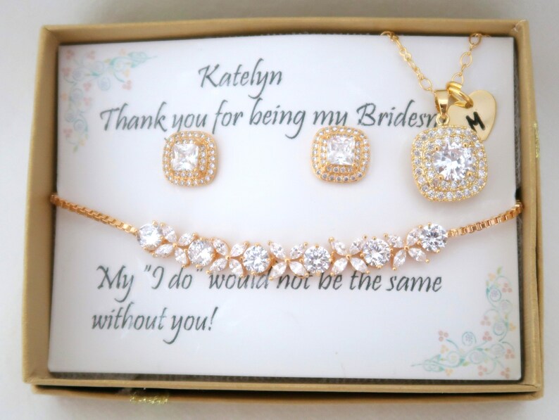 K&ouml;nnte beinhalten: Ein goldfarbenes Schmuckset, bestehend aus einer Halskette, Ohrringen und einem Armband, pr&auml;sentiert in einer Geschenkbox. Die Ohrringe und die Halskette haben quadratische, klare Edelsteine. Das Armband hat ein florales Design.