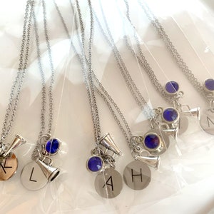 May include: Silver-toned necklaces with a chain, a blue gem, a megaphone charm, and a round charm with a letter engraved on it. The necklaces are displayed on a white surface.