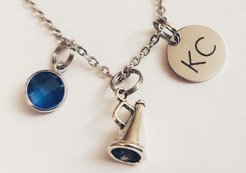 May include: Silver-toned necklace with three charms: a blue gemstone, a megaphone, and a round pendant engraved with "KC". The necklace is on a white background. A piece of jewelry.
