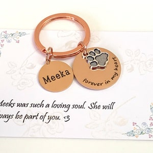 May include: A rose gold keychain with two circular charms. One charm is engraved with the name "Meeka", the other with a paw print and the words "forever in my heart". A card with floral accents and the text "Meeks was such a loving soul. She will always be part of you. <3" is in the background.
