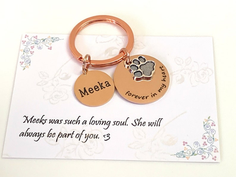 May include: A rose gold keychain with two circular charms. One charm is engraved with the name "Meeka", the other with a paw print and the words "forever in my heart". A card with floral accents and the text "Meeks was such a loving soul. She will always be part of you. <3" is in the background.
