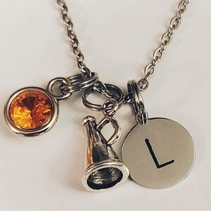 May include: Silver-toned necklace with multiple charms. Charms include a round orange gemstone, a megaphone, and a circular pendant with the letter "L". The necklace is on a white background.